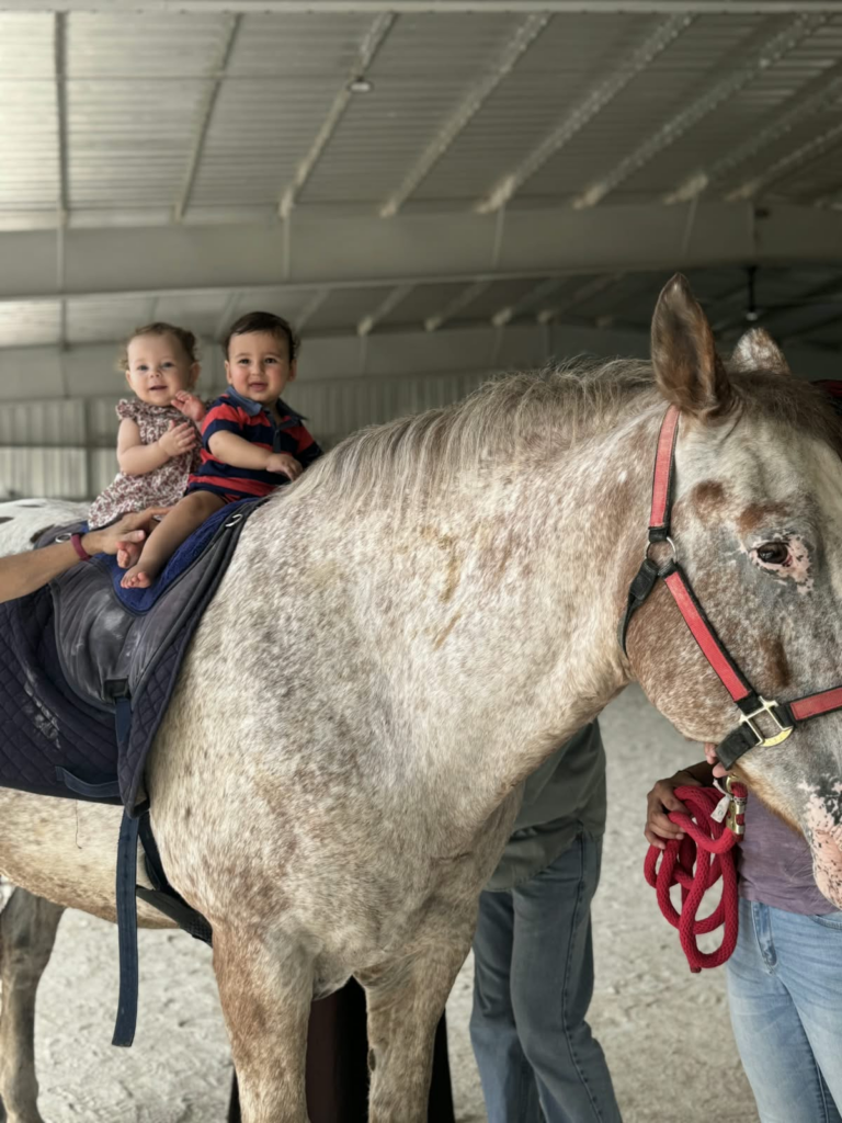 Dos bebés sonrientes montados en un caballo manchado, sostenidos por la mano de un adulto
